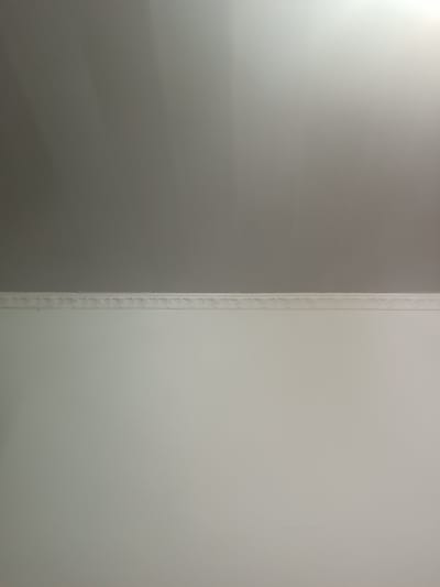 The image depicts a close-up, indoor view of a wall and ceiling, divided by a decorative cornice. The lower portion of the image, occupying approximately two-thirds of the frame, shows a plain, white painted wall. Above this, a decorative white cornice runs horizontally across the image, featuring a repeating, intricate pattern. The upper portion of the image shows a ceiling, which appears to be a light gray color, transitioning to a lighter, brighter area towards the right side, suggesting a light source from that direction. The surface of the ceiling has a subtle, textured pattern, possibly from paint or wallpaper. There are no people or identifiable objects other than the architectural elements of the room. The scene appears to be a generic interior, likely a room in a residential or commercial building, with no specific or discernible details that would associate it with the city of Бидайкөл, Kazakhstan.