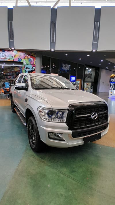 A silver pickup truck is parked indoors, possibly in a shopping mall. The truck is facing the camera, angled slightly to the left. It has a black grille with a chrome emblem and black rims on the wheels. The headlights are illuminated with blue accents. In the background, there are store displays with colorful graffiti art on the walls. The floor is a mix of green and orange-brown colors. A person is visible sitting at a table to the left of the frame, appearing to be in a cafe or food court area. The overall environment suggests an indoor commercial space. The location is described as Unknown, Venezuela.