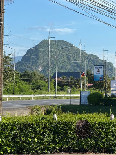 This photograph depicts a roadside scene in Ban Ko Pru, Thailand. A two-lane road runs vertically through the image, flanked by lush green vegetation on the right side. A mountain covered in dense trees provides a backdrop in the distance, with power lines stretching across the scene. Signs are visible alongside the road, including one advertising "เตี๋ยวตุ้น" (Tiew Tun), a noodle dish, with its opening hours listed. Additional signs and smaller advertisements are also present. The sky is clear with a light blue hue. The overall scene suggests a rural or semi-rural setting, with the road possibly leading through or past Ban Ko Pru.
