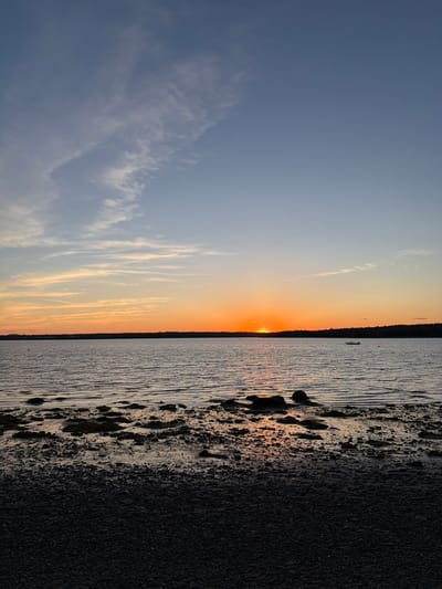 The image shows a sunset over the water, possibly taken at a beach near Bar Harbor, Maine, United States. The sky transitions from a clear blue to shades of orange and yellow near the horizon, where the sun is setting. Long, thin clouds are visible in the sky. The shoreline consists of a dark, gravelly beach bordering the water. The water reflects the colors of the sunset.