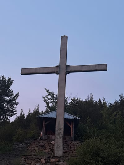 The image depicts a large, metal cross standing on a rocky, elevated area surrounded by trees. The cross is tall and appears to be made of industrial-style metal beams. At the base of the cross, there is a small, open-sided shelter with a metal roof, supported by wooden beams. The shelter is built on a stone foundation. The ground in front of the shelter is also covered in stones. A person can be partially seen inside the shelter. The sky above is a pale blue, transitioning to a soft purple hue near the horizon, suggesting either dawn or dusk. The surrounding environment is a dense forest, with various types of trees and foliage. The location is identified as being in the city of Strazha, Bulgaria.