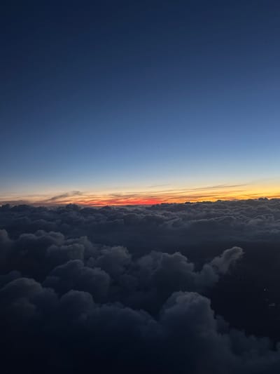The image displays an aerial view captured from above a substantial cloud layer during either sunrise or sunset. The upper approximately 60% of the frame features a sky transitioning from a deep blue at the top, fading to a lighter blue, and then to a prominent horizontal band of vibrant orange, red, and yellow light along the horizon. This illuminated band sits directly above a dense, undulating expanse of dark gray clouds that fills the lower 40% of the image. The cloud tops exhibit varying degrees of light and shadow, appearing darker in their deeper sections. In the bottom right corner, through a discernible break in the cloud cover, faint clusters of artificial ground lights are visible, indicating a populated area below. Two minute, unidentified dark specks are present in the upper blue sky. No individuals, specific structures, or discernible actions are visible. The visual content does not provide sufficient information to confirm the specific ground location as Santiago Cuautlalpan, Mexico.