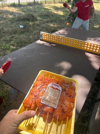 This media file shows an outdoor scene with a person playing table tennis and a tray of raw chicken skewers. The person, wearing a red t-shirt and light-colored pants, is in the process of hitting a table tennis ball. A red table tennis paddle is visible in their hand, and a small orange ball is in the air.

To the right of the table, a yellow plastic tray holds numerous chicken skewers, seasoned and ready for cooking. A label on the packaging displays text in Bulgarian, including "ЕРКОМЕС," which appears to be a brand name, and information about the product being "chicken thigh skewers without bone." There is also a weight of "1,000 kg" indicated.

The setting appears to be a park or a yard, with green grass, trees casting shadows, and sunlight suggesting a daytime activity. The overall mood is casual and recreational, with the focus on both a sport and preparing for a meal, possibly a barbecue. The presence of the table tennis equipment and the prepared food hints at a social gathering or a leisurely afternoon outdoors.