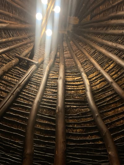 The image displays an upward-angled view of the interior ceiling of a circular or conical structure, located in Dulovine, Montenegro. The ceiling features a traditional thatched design, with layers of golden-brown straw or similar plant material laid between dark, roughly hewn wooden beams that radiate from a central point. Approximately four visible spherical light bulbs, emitting bright white light and producing prominent lens flares, are mounted near the apex of the ceiling, providing illumination to the interior. No individuals or specific actions are discernible within the frame.