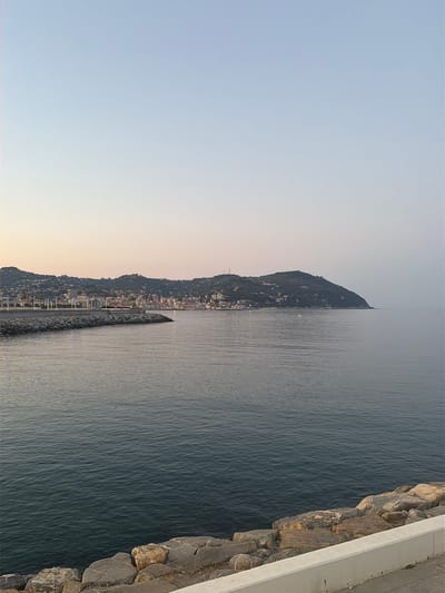 This is a landscape photograph taken from a waterfront promenade, looking out over a body of water, likely the Ligurian Sea. The image is oriented vertically, with the sky at the left and the sea and coastline to the right.
The left side of the image shows a pale, gradient sky, transitioning from a soft blue at the top to a warm, peachy hue near the horizon. This suggests it is either sunrise or sunset. The right side of the image is dominated by the deep blue water, with subtle ripples indicating a calm sea.
Along the distant coastline, a densely built town is visible nestled against a steep, tree-covered hillside. The buildings appear to be clustered together, with some lights twinkling, further supporting the idea of dawn or dusk. A breakwater, constructed of large stones, extends from the shoreline into the sea, creating a segmented line in the water. In the water, a small white boat with its lights on is visible.
In the foreground on the far right, there is a concrete walkway or promenade with a rocky embankment bordering the sea. The rocks are varied in size and color, with rough textures. The overall mood of the image is serene and tranquil, capturing a peaceful coastal scene at the cusp of day. There is no visible text in the image.
