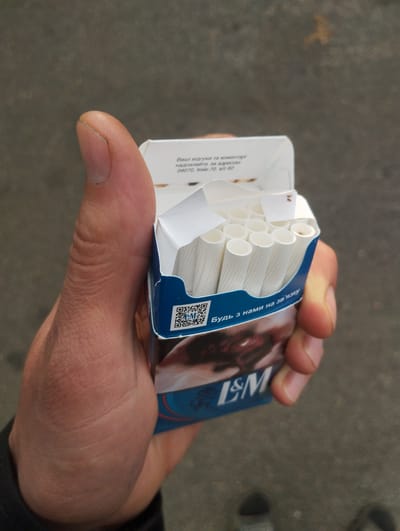 A person's left hand is holding an open pack of L&M cigarettes. The pack is blue and white with text in Ukrainian. There are several cigarettes visible inside the pack. The background appears to be an outdoor, urban setting, possibly a street or sidewalk. There is no specific indication of the location being Vinkivtsi, Ukraine.