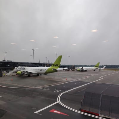This image presents an overcast view of an airport tarmac in Skulte, Latvia, observed from an elevated position, likely within a terminal building.

In the immediate foreground, a dark gray tarmac surface is visible, marked with white lines, including a prominent double white line that curves from the bottom right towards the center, and dashed white lines. A large red triangular marking with a white interior is painted on the tarmac in the bottom left. Circular white markings are also present on the tarmac. To the far right, a section of a dark metal mesh fence with a red base is partially visible. The tarmac shows signs of dampness, indicating recent rain or high humidity.

In the midground, two white AirBaltic aircraft are parked on the tarmac. The aircraft on the left, registered YL-ABA, features a distinctive lime green tail with "Baltic" written in white, and lime green engine cowlings. Passenger stairs are connected to its front left door, and two ground personnel in high-visibility yellow jackets are standing near the base of the stairs. Orange traffic cones are placed around this aircraft. A dark vehicle is partially visible behind the ground crew. The aircraft on the right, registered YL-ABC, also displays the AirBaltic livery with a lime green tail. It is connected to a passenger boarding bridge labeled "103," which also bears "Revolut" branding. A smaller ground service vehicle is positioned near the nose of this aircraft.

In the background, a modern airport terminal building with dark paneling and large windows spans across the scene. Several tall floodlight poles are positioned between the aircraft and the terminal. Various antennas and structures are visible on the terminal's roof. The word "Revolut" is also visible on the terminal building itself. Beyond the airport infrastructure, a dense line of dark green trees forms the horizon under a uniformly gray, heavily overcast sky. The upper portion of the image reveals the interior ceiling or window frame of the viewing area, with several square, warm-toned lights or reflections scattered across the otherwise plain gray surface.