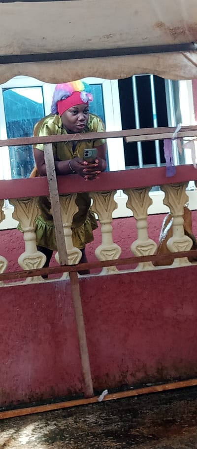 A dark-skinned adult female is depicted leaning forward over a balcony railing in the city of Udi, Nigeria. She wears a greenish-gold short-sleeved garment with a ruffled hem and a red head wrap adorned with a colorful, multi-striped fluffy fabric piece. She holds a dark-colored smartphone horizontally with both hands, positioned below the railing, and her gaze is directed slightly upwards and to the viewer's left. The railing consists of a reddish-brown wooden top rail, cream-colored decorative balusters, and a lower reddish-brown horizontal bar, all mounted on a red-painted wall. A raw wooden beam extends vertically, partially obscuring the woman. Behind her, two windows are visible; the left window reflects a blue tint, while the right window shows dark vertical elements. Light-colored decorative strips hang from a structure on the right side of the background. A light beige canopy or ceiling is visible at the top of the frame. The area below the balcony appears to be a dark, textured ground surface.