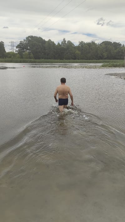An adult male, wearing dark blue swimming trunks, walks away from the camera into a body of water. He is shirtless and his back is to the viewer. The water is shallow around his knees, creating ripples and splashing as he moves forward. The background shows a calm lake or river with a treeline on the far bank. Power lines are visible crisscrossing the sky above the trees. The sky is overcast. The scene appears to be a natural outdoor setting, possibly a lake or riverbank, and not specifically within the city of Ramon, Russia.