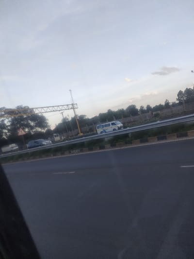 The scene depicts a multi-lane highway in Theta ward, Kenya, observed from a low angle, likely from a vehicle. In the immediate foreground, a dark asphalt road with faint white lane markings is visible, bordered by a black and tan checkered curb. A dark, indeterminate object partially obscures the lower-left quadrant. Beyond the foreground, a metal guardrail separates traffic lanes. On the opposing side of the guardrail, three vehicles are present: a white minibus with blue accents, a dark sport utility vehicle (SUV), and a white light commercial truck. An overhead yellow metal gantry structure with a vertical support pole is positioned above the road on the left. A triangular warning sign, partially obscured, is visible to the left of the gantry. The highway is flanked by dense green trees, behind which a light-colored concrete wall or fence extends. The sky is overcast with scattered clouds, suggesting daylight conditions. No human figures are discernible.