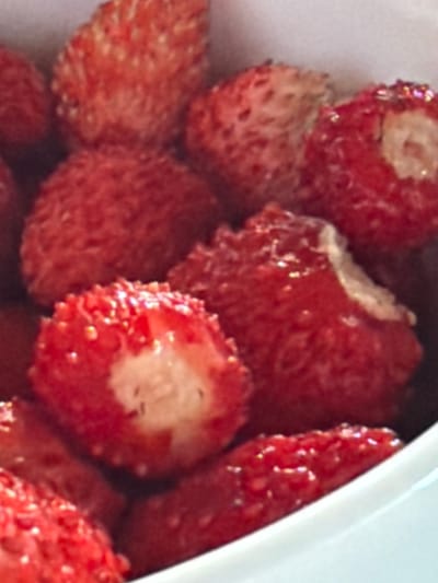 This media file is a close-up, eye-level shot of several halved strawberries, likely wild strawberries given their size and texture. The strawberries are red and appear ripe, with visible seeds on their surface. Some of the strawberries are cut in half, revealing their white interior flesh and slightly darker red outer layer. The texture of the strawberries is rough and textured, suggesting they are fresh. The lighting is bright and appears to be natural, casting a slight sheen on the berries. The background is mostly out of focus, but appears to be a white container or surface, possibly a bowl or plate, and the edge of another white object is visible on the right side, which could be the side of the container or another dish. The focus is entirely on the strawberries, highlighting their natural beauty and texture. There is no visible text in the image, nor any people or animals. The overall impression is one of fresh, natural produce. The context of Aespa, Estonia, suggests these might be locally sourced berries, possibly enjoyed as a snack or dessert.