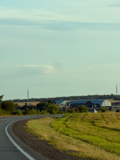 The image depicts a scenic rural landscape near Togashevo, Russia, under a pale blue sky with wispy clouds. The foreground is dominated by a curved asphalt road with white lane markings, transitioning into a grassy embankment. In the midground, a line of trees separates the road from a cluster of buildings, including what appear to be agricultural structures with white, arched roofs and other residential or utility buildings. Power lines and transmission towers are visible in the distance, suggesting the presence of infrastructure in the area. A few vehicles are present, including a white SUV and a dark-colored car, suggesting light traffic. The overall impression is of a peaceful, functional rural setting.