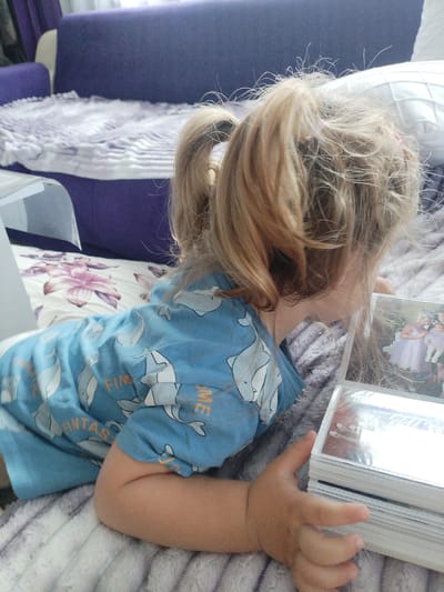 A young child with blonde hair in pigtails is lying on a bed, looking at a stack of photographs. The child is wearing a blue t-shirt with a pattern of grey dolphins. The bed is covered with a grey and white striped blanket, and a floral-patterned pillow is visible. In the background, a purple couch is also visible. The scene appears to be indoors, likely a bedroom or living room. This image was taken in Rakitovo, Bulgaria.