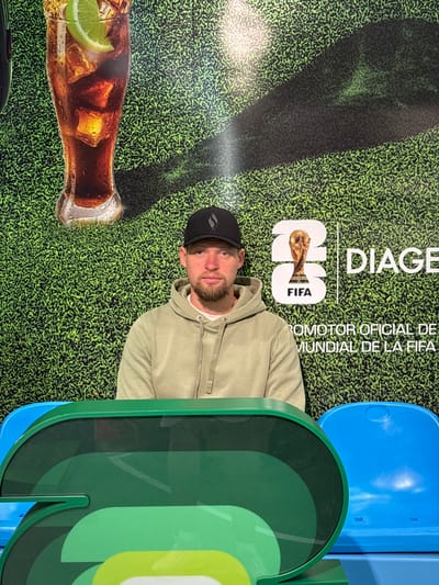 This media file shows a man wearing a light green hoodie and a black baseball cap, posing in front of a large backdrop. The backdrop features a green, textured design resembling grass. To the left of the man, a close-up of a glass bottle filled with a dark beverage and ice cubes, garnished with a lime wedge, is visible.
Dominating the background are large, stylized green letters that appear to be part of a logo or branding. To the left of these letters, a graphic displays the FIFA World Cup trophy and the text "DIAGE" and "FIFA". Further to the left, the text "POMOTOR OFICIAL DE MUNDIAL DE LA FIFA" is present. A bright blue object, possibly seating, is visible in the upper right corner.
The man is looking towards the camera with a neutral expression. The lighting suggests an indoor setting with artificial light, as indicated by the shadows cast on the backdrop. The overall scene appears to be promotional, possibly related to a sporting event or a beverage brand, given the presence of the FIFA logo and the Coca-Cola-like imagery. The location context is Ezeiza, Argentina.