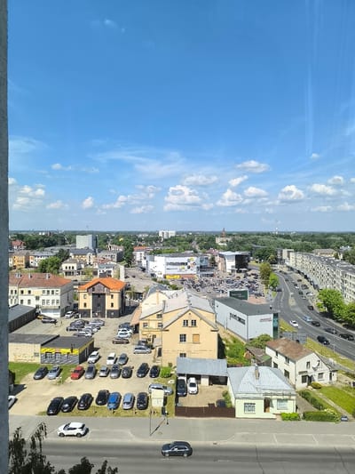 The image provides an aerial view of a section of the city of Panevėžys, Lithuania, on a bright, sunny day with a blue sky dotted with white clouds. The scene depicts a mix of residential and commercial buildings, a busy road with traffic, and several parking lots filled with cars.
In the foreground, a gray building with a textured surface is partially visible on the left, suggesting it's a taller structure from which the photo was taken. Below this, a road with a white painted line marking parking spaces runs horizontally. A white SUV is parked in one of these spaces. Further down the road, a dark colored sedan is visible.
Moving further into the scene, the landscape is characterized by a variety of buildings. Some are multi-story residential blocks with light-colored facades, while others are smaller, older houses with tiled roofs. Commercial buildings are also present, including a modern, gray building with large windows and a sign that appears to be a business. Adjacent to it, a yellow building with a black facade and a red-tiled roof stands out.
A significant feature of the image is a moderately busy road that curves through the scene. Numerous cars are seen driving on this road, indicating active traffic flow. Several parking lots are visible, densely packed with cars of various colors and models. One parking lot is situated in front of a yellow and gray building, and another is near a larger commercial complex.
The background of the image reveals more of the city, with a dense concentration of trees interspersed with buildings of varying heights. In the far distance, taller structures, possibly industrial chimneys or taller buildings, are visible against the horizon. The overall impression is that of a functional urban environment on a clear summer day.