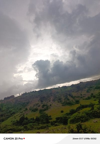 This image, taken in Nguroje, Nigeria, captures a dramatic sky filled with heavy, dark clouds that obscure much of the sun, creating a somber mood. A sliver of bright light breaks through the clouds on the left side of the frame. The lower portion of the image shows a lush, green, and hilly landscape. Vegetation, including trees and shrubs, covers the slopes, with patches of grass and possibly some cultivation. The terrain appears rugged and uneven. The bottom of the image contains a white overlay with text: "CAMON 20 Pro" on the left and "24mm f/1.7 1/598s ISO52" on the right. There are no people or animals visible in the scene. The overall impression is one of natural, undeveloped scenery under a brooding sky.