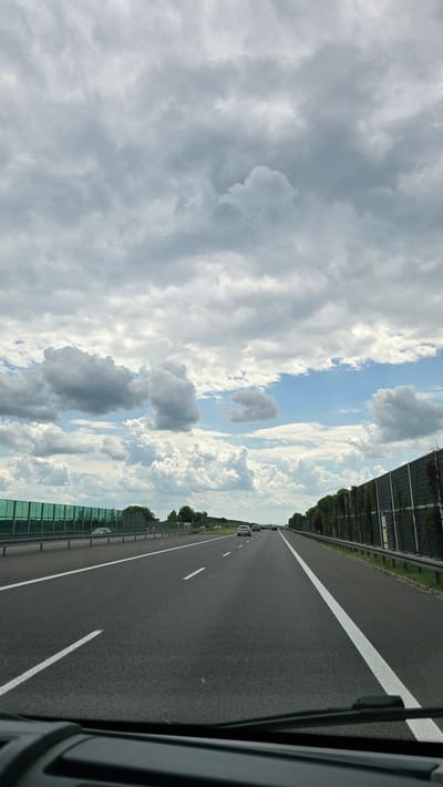 The image shows a view from a car driving on a highway in the city of Świerklany, Poland. The sky is filled with a dramatic display of clouds, ranging from dark, heavy masses to lighter, wispy formations, with patches of blue sky visible. The highway is paved asphalt with clear white lane markings. On the left side of the highway, there is a green sound barrier. On the right side, there is a similar sound barrier covered in ivy. Several cars are driving in the same direction as the viewer. The overall scene suggests travel on a partly cloudy day.