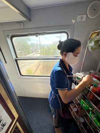 A woman wearing a blue uniform and a white face mask is seen on a train, likely traveling through Sơn Tịnh, Vietnam. She is standing next to a shelf filled with various products, including drinks and snacks. Sunlight streams through the train window, illuminating the scene and casting shadows on the interior. The woman is holding a white cup with a green label and appears to be attending to the merchandise. The train's interior is utilitarian, with grey walls and dark speckled flooring. Outside the window, lush green trees and a dusty road are visible, characteristic of a rural Vietnamese landscape. The presence of a train suggests a mode of transportation common in Vietnam, and the surrounding vegetation points to a location outside of a dense urban center.