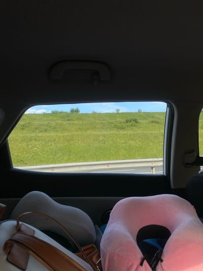 The image is taken from inside a car, looking out the rear window. The view outside is of a grassy hillside under a blue sky with scattered white clouds. A metal guardrail runs horizontally across the lower part of the view, suggesting the car is on a road. Inside the car, in the foreground, are two travel pillows, one pink and one grey, and the top of a white bag with brown straps. The overall impression is of a journey or travel. The location, "Пятый километр, Russia," is a street name or a marker indicating a distance from a central point, often found along roads or in rural areas, and does not specify a particular city, but rather a location within Russia.