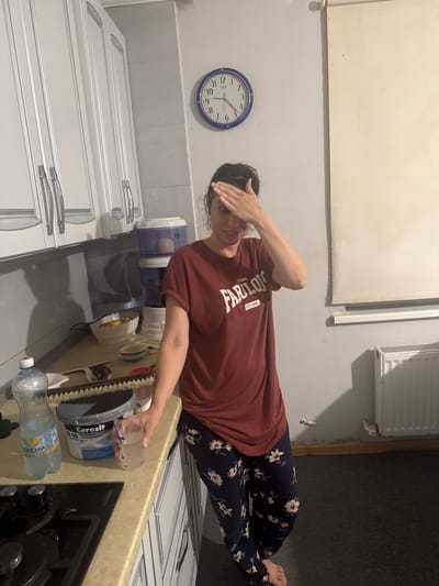The image shows a woman in a kitchen with white cabinets and a beige countertop. She is wearing a maroon t-shirt with the word "FABULOUS" printed on it and dark blue floral patterned pants. Her right hand is on her forehead. On the countertop, there is a bottle of water, a container labeled "Ceresit CE 40", a white bowl with oranges, and a glass of clear liquid. A water dispenser with two blue tanks is visible behind the woman. A clock with a blue frame is on the wall, showing approximately 1:55. A white radiator is mounted on the wall to the right of the woman. The scene appears to be indoors, possibly in a residential kitchen. No specific location in Novosiltsi, Ukraine can be determined from the image alone.