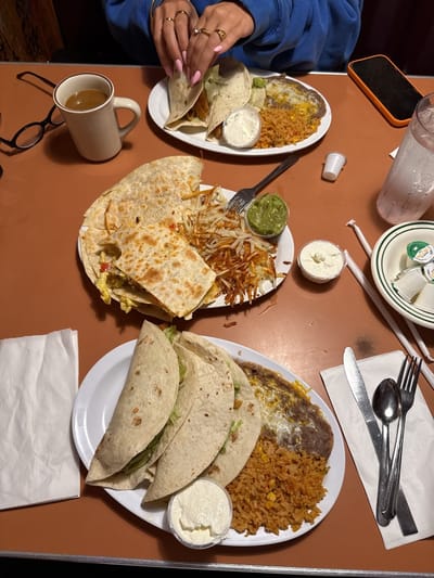 The image shows a table at a restaurant in Huntington Beach, California. Three plates of food are visible: two plates with three tacos each, rice, beans, and a small container of sour cream; and one plate with a quesadilla, hash browns, and guacamole. A mug of coffee, a glass of water, a cell phone, a napkin, and silverware are also on the table. One person is visible with their hands on the table near one of the taco plates. They are wearing rings.