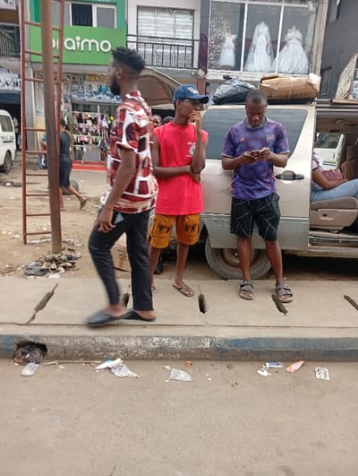 A street scene captures three men standing on a sidewalk beside a parked van. One man in a patterned shirt walks by, while another in a red tank top and blue baseball cap stands with his arms crossed. The third man, wearing a purple shirt, focuses on his phone. Trash litters the curb and street. Behind them, shops display goods, including a wedding dress in a window. A ladder leans against a building in the background.