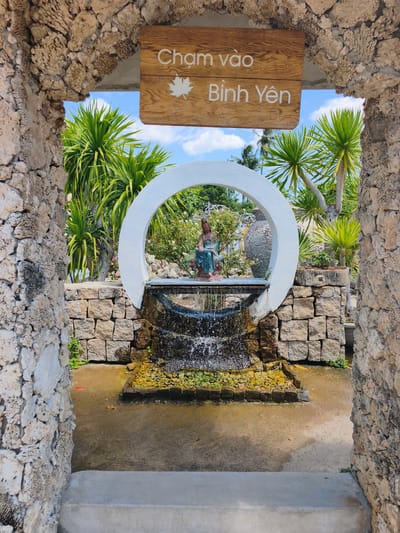 The image captures a view through a stone archway, leading to a tranquil garden scene in Tuy An Nam, Vietnam. A wooden sign above the arch reads "Cham vào Bình Yên" with a white maple leaf symbol. Inside, a circular white structure frames a small waterfall cascading over rocks. A statue of a serene figure, likely a deity or a meditating person, is seated behind the waterfall. Lush green palm trees and other vegetation are visible in the background under a bright blue sky with scattered clouds. The ground in front of the waterfall is wet, indicating the presence of water. The overall scene evokes a sense of peace and natural beauty.