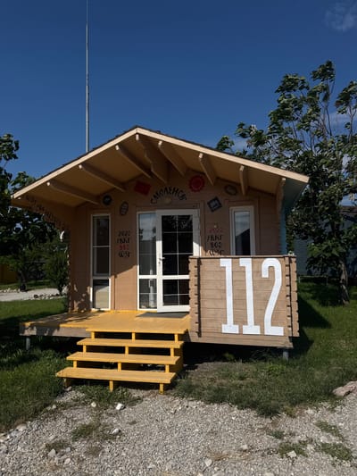 A small, wooden cabin with a yellow porch and stairs is pictured under a clear blue sky. The cabin has a light brown wooden exterior and a tan roof. White numbers "112" are prominently displayed on the front right side of the cabin. The cabin is situated in a grassy area with gravel in the foreground. Trees are visible on the right side of the image. The cabin is located in the city of Sudak, Ukraine.