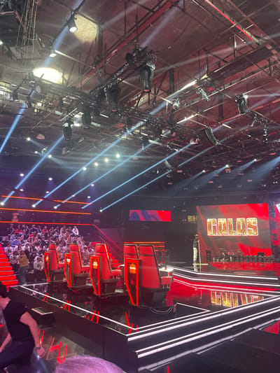A rotated stage with lighting rigs and a large screen reading "THE VOICE" in glittery gold letters is visible. The stage setup features multiple red chairs and geometric structures outlined in bright white lights. A crowd of people is seen seated below, illuminated by spotlights. The interior architecture includes a high, industrial-style ceiling with exposed beams and rigging.