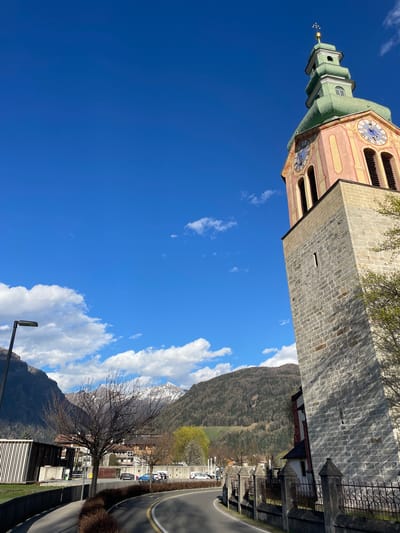 The image depicts the Twelve Apostle Church (Zwölferturm) in Sterzing - Vipiteno, Italy, seen from a low angle looking upwards. The tower's architectural details, including its clock faces and pointed spire, are visible against a bright blue sky with scattered clouds. The mountains and some snow-covered peaks in the background blend in with clouds. The view also captures sections of the street and nearby buildings. A barren tree stands along the street, and a lamp post is on the side.