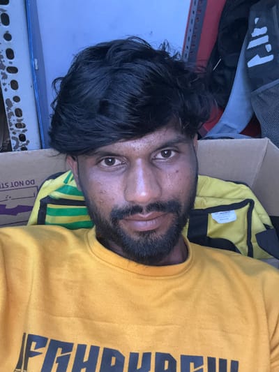 The image depicts an adult male in an indoor setting within Palghar, India. The subject, positioned centrally and looking directly at the viewer, has dark skin, dark hair, and a full beard. He is wearing a yellow long-sleeved sweatshirt with dark lettering partially visible across the chest. His eyes are dark brown.

The background elements include a cardboard box to the subject's left and behind him, displaying partially visible text "DO NOT STO[P]". Behind his right shoulder, a yellow and green striped fabric item, possibly a bag, is visible, with a white tag attached that reads "Style to Style Out", "www.luffers.in", "info@luffers.in", and "Made in India". Further in the background, to the left, is a grey metal shelving unit with multiple circular perforations, alongside a blue-painted wall. To the right, another metal rack, possibly red, and a dark grey backpack with white markings are visible. The overall lighting appears dim.