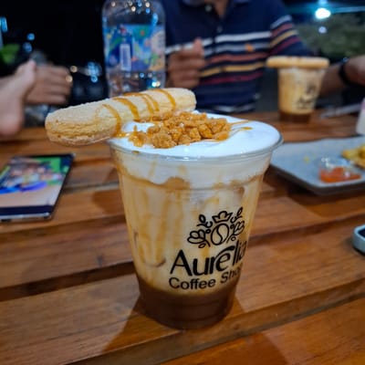 The image captures a close-up view of a caramel iced coffee drink, presented in a clear plastic cup with the "Aurelia Coffee Shop" logo printed on it. The drink is topped with a thick layer of white foam, sprinkled with crushed biscuits and drizzled with caramel sauce. A single, elongated biscuit, also coated in caramel, is artfully placed across the foam. The cup sits on a wooden table, suggesting an outdoor or casual indoor setting. In the background, slightly out of focus, a person wearing a striped shirt and another person's hand are visible, along with a bottle of water. A smartphone displaying a colorful image is positioned to the left of the coffee cup on the same table. The overall lighting suggests an evening or dimly lit environment, possibly a cafe. The presence of the "Aurelia Coffee Shop" logo indicates the drink originates from this establishment. While the specific location within Mesjid Tuha, Indonesia, is not directly depicted, the context of a coffee shop implies a commercial establishment serving patrons.