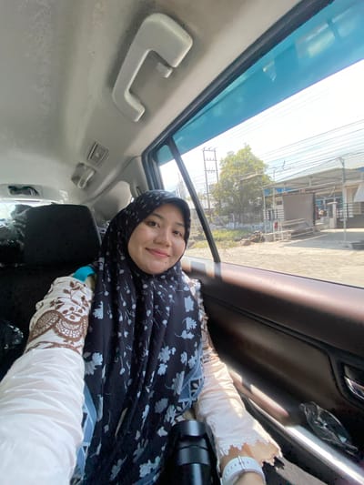In Sungai Lakum, Indonesia, a woman wearing a dark hijab with a floral pattern and a white long-sleeved shirt with a brown lace design is captured in a selfie from inside a vehicle. She is seated on the right side of the back seat, her face illuminated by sunlight filtering through the car window. The interior of the car is visible, including the ceiling, a grab handle, and the dark brown door panel. Outside the window, a street scene unfolds with buildings, trees, and power lines under a clear sky. The woman is smiling, suggesting a candid or posed moment of travel.