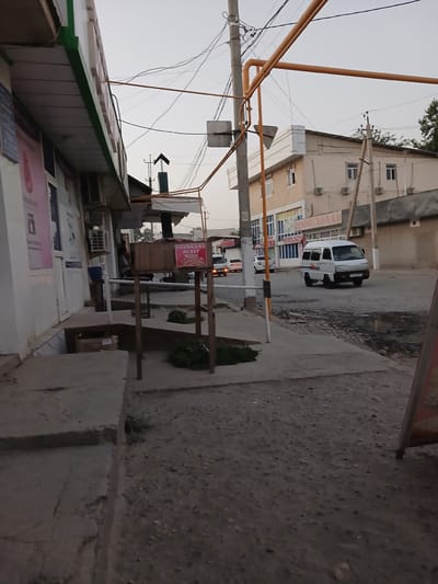 The image captures a street scene in Denov, Uzbekistan. On the left, a storefront with a white facade and a green trim is visible. Above the storefront, a sign with blurry text is mounted. Part of a pink advertisement poster is visible on the left side of the storefront. Further down the street, a wooden structure with a sign that reads "TOSHKENT SUTLI NON" (Tashkent Milk Bread) is present, displaying some items on top. The street itself is paved with asphalt and shows signs of wear and tear, with puddles and debris. A white minivan with a license plate is driving on the street, and other vehicles are visible in the background. Buildings line the opposite side of the street, including a light-colored building with multiple windows and air conditioning units. Overhead, a complex network of electrical wires and yellow pipes crisscrosses the sky, supported by utility poles. The sky is overcast. The scene appears to be captured in the late afternoon or early evening.