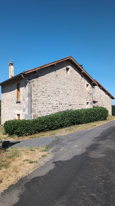 A side view of a stone house with a wooden-shingled roof is captured under a clear blue sky. The house has a chimney with an antenna on top and several small windows. A dense green hedge runs along the base of the house, separating it from a gravel path and a paved road. The surrounding landscape is dry and appears to be a rural area. The location is identified as Sainte-Sigolène, France.