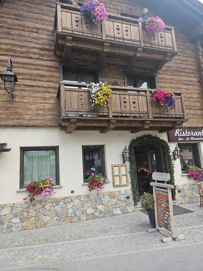 This is an eye-level, medium shot of the exterior of a rustic wooden building in Livigno, Italy, likely a restaurant or hotel. The building features a traditional alpine architectural style with prominent wooden beams and balconies adorned with colorful hanging flower baskets. Two balconies are visible, each with wooden railings featuring heart-shaped cutouts. The upper balcony has two individuals visible, one leaning on the railing and looking to the left, the other partially obscured. Flower baskets with pink, purple, and yellow flowers hang from both balconies. The lower portion of the building is a cream-colored stucco with stone accents at the base. Two ground-floor windows are visible, also with flower baskets below them. An arched entryway is decorated with evergreen garlands and features two lantern-style light fixtures on either side. A wooden signpost stands in front of the entryway, displaying a menu for "Ristorante con terrazzo" and listing the establishment as "La Montanina - Bar, Ristorante, Hotel." The ground in front of the building is paved with cobblestones.