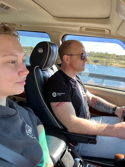 Two people are seated in a car. The location is indicated by the word "КОЛЬСКАЯ АЭС" on the driver's sleeve, which translates to Kola Nuclear Power Plant, in the Murmansk region of Russia, in the city of Monchegorsk. A man with sunglasses sits in the driver's seat, gazing out the window. He has a watch and a tattoo on his right arm. He is wearing a black shirt and a seatbelt. His head is resting on the headrest. Another person, possibly a woman, sits in the passenger seat. They are wearing a dark-colored hoodie, secured with a seatbelt. They are also gazing out the window. The car is moving past a body of water with trees visible in the background. The interior of the car appears to be well-maintained.