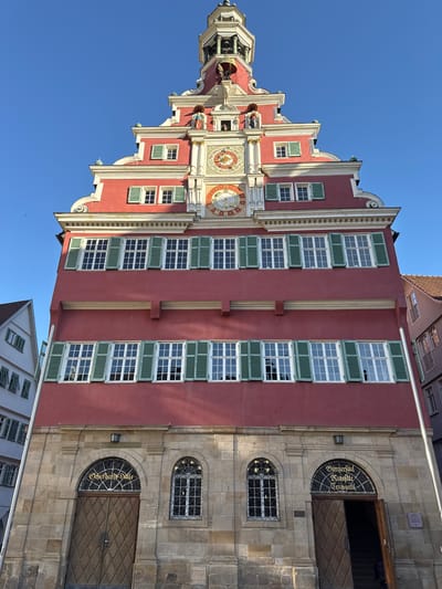 The image shows the Esslingen am Neckar town hall, known as the Esslinger Rathaus, situated in Esslingen am Neckar, Germany. The building is a prominent, multi-story structure with a distinctive red facade, a tiered roofline, and numerous windows with green shutters. The architectural style appears to be historic, possibly Renaissance or Baroque.

At the top of the building, a bell tower with a spire is visible against a clear blue sky. Below the tower, there are decorative figures and two large clock faces. The main body of the building features rows of windows, with each window divided into multiple panes. The lower section of the building is constructed of stone blocks and features large wooden doors and arched windows. Golden lettering is visible above the doors.

In the foreground, the main entrance to the town hall is open, revealing a dark interior. Two individuals are partially visible entering or exiting the building. To the left and right of the town hall, other buildings are partially visible, suggesting an urban setting. The overall scene depicts a well-preserved historic building under a bright, sunny sky.