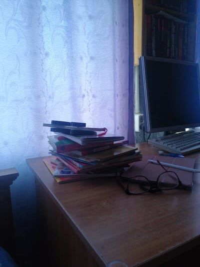 The image depicts a desk in an indoor setting, likely a home office or study, in Dubrovka, Russia. On the desk, a stack of books is prominently displayed, leaning against a patterned curtain. To the right, a computer monitor sits atop a keyboard, suggesting a workspace. Adjacent to the books, a pair of eyeglasses rests on a tablet, and a stylus lies nearby. The overall ambiance is one of quiet study or work, with the natural light filtering through the curtains casting a soft illumination on the scene. The presence of books, a computer, and writing implements indicates an environment conducive to learning or professional tasks. The background shows a bookshelf filled with books.