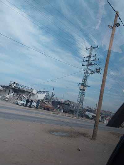 The image portrays a scene of extensive urban destruction in a-Zawayda, Palestinian Territory, under a sky exhibiting light blue areas with wispy white cloud formations. The foreground consists of a dirt and gravel surface, containing a small puddle. A paved road traverses the midground horizontally. On the right, a vertical wooden utility pole features a single light fixture. Multiple overhead power lines extend across the upper portion of the frame. To the left of the road, a white sedan is parked amidst significant piles of concrete rubble and the partially collapsed remains of multi-story buildings. Approximately three to four individuals, identified as adult males in dark clothing, are standing near the white sedan. Further to the right, a flatbed truck laden with debris is present. A prominent, leaning metal lattice power transmission tower, equipped with multiple cross-arms and insulators, occupies the mid-right of the scene. The background reveals additional severely damaged structures and debris stretching towards the horizon.