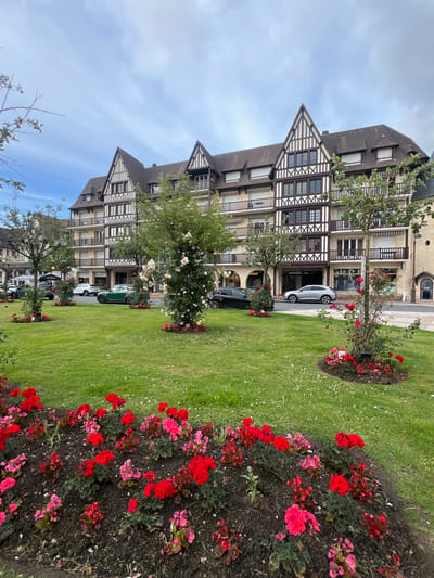 This is a photograph of a building in Deauville, France. The building is a multi-story structure with a distinctive half-timbered facade, featuring dark wood beams against a light-colored exterior. The architecture is characteristic of traditional French coastal towns. In the foreground, a well-maintained lawn is dotted with small white flowers. Several flower beds are visible, filled with vibrant red and pink flowers, most prominently geraniums. There are also rose bushes, one of which is adorned with white blossoms, adding to the floral display. Trees are scattered throughout the lawn, some with fuller foliage and others with delicate branches. Several cars are parked on the street in front of the building, including a green Mini Cooper, a silver SUV, and a black hatchback. The sky is overcast with a mix of blue sky and gray clouds. The scene appears to be a public space, likely a park or square in front of shops or residences in Deauville.
