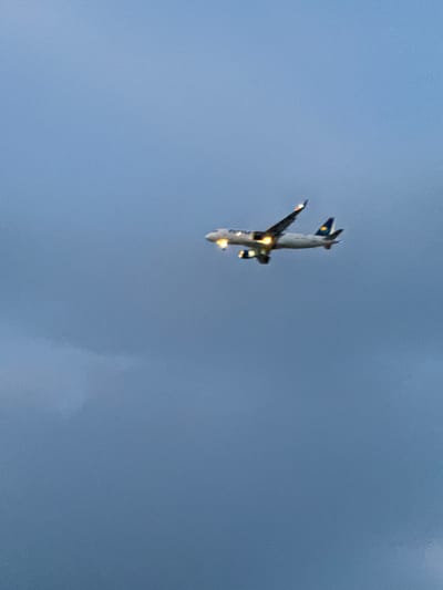 An airplane is in flight against a cloudy sky. The plane is white with yellow lights illuminating its engines and windows. A yellow and blue logo is visible on the tail of the airplane. The airplane is positioned in the upper right quadrant of the image. The sky is a gradient of blue and gray, indicating it is likely dusk or dawn. There are no buildings, people, or other objects visible in the frame. The image was captured in Grenay, France.