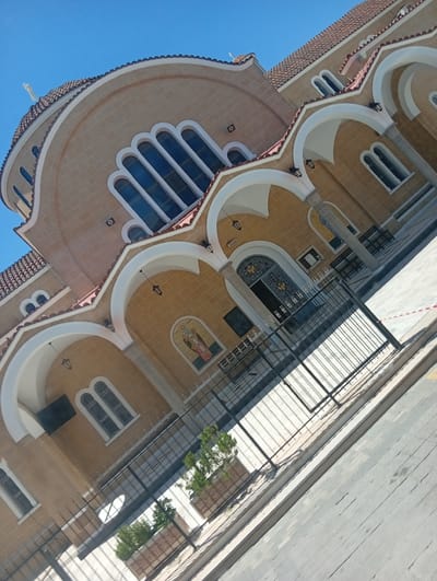 The image captures a vibrant exterior view of a large, light-colored church or cathedral, likely an Orthodox church, under a clear, bright blue sky. The building is constructed from light brown or tan stone/stucco, topped with red-tiled roofs and a prominent central dome featuring a white cross.
The architectural design is characterized by numerous white arched porticos and window frames, creating a striking contrast with the main façade. Multiple windows, some tall with arched tops and others smaller, are visible throughout the structure. A main entrance features ornate black metal double doors with decorative patterns, possibly religious symbols. Above one of the arched entrances, a colorful mosaic depicts a winged saint or angel holding a staff. Another smaller mosaic icon is also visible near the main doors. Lanterns or light fixtures hang beneath some of the arches.
In the foreground, a black metal fence with vertical bars separates the church grounds from a paved sidewalk and street. Strong, distinct shadows are cast by the fence and parts of the building, indicating bright sunlight and suggesting the photograph was taken on a clear, sunny day, likely late morning or early afternoon. A small potted plant adds a touch of greenery near the fence. The perspective is tilted, giving the image a dynamic, upward-looking composition. The architectural style, prominent dome, and mosaics are consistent with churches found in Paralimni, Cyprus. No people are visible, and no legible text can be identified in the image.