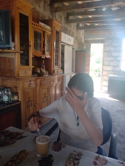 In Nor Geghi, Armenia, within a rustic, stone-walled room with exposed wooden beams, a person is seated at a table. The person, whose face is partially obscured by their hand, wears a white, short-sleeved, ribbed top and a dark beaded bracelet on their left wrist. They are stirring a light brown beverage in a clear glass with a spoon. The table is covered with a patterned cloth, and a variety of nuts, possibly pistachios and almonds, are scattered across it. To the left of the table, a tall, wooden cabinet with glass-fronted doors and drawers is visible, containing various items. In the background, an open doorway reveals a sunlit outdoor scene with greenery. The overall environment suggests a casual, possibly home setting.