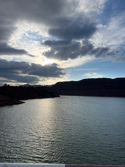 The image shows a landscape in Tabaroucht, Morocco, featuring a large body of water, possibly a lake or reservoir. A dark, rocky mountain or cliff formation is situated alongside the water. The sky is partially cloudy with visible sunlight piercing through the clouds. The water has a rippled surface reflecting light, giving it a shimmering appearance. A structure or part of a bridge is visible on the right side of the image.