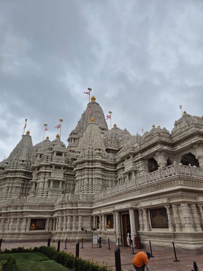 The image captures the exterior of the BAPS Shri Swaminarayan Mandir in Robbinsville Township, New Jersey, United States of America. The temple, constructed primarily of white stone, features intricate carvings and multiple tiered roofs topped with golden spires and flags. The sky above is overcast with a mix of light and dark gray clouds. In the foreground, a person wearing an orange shirt and a blue cap is kneeling and appearing to clean the ground. Black stanchions with chains are set up along the pathway in front of the temple. Green grass and some low-lying bushes are visible to the left of the frame.