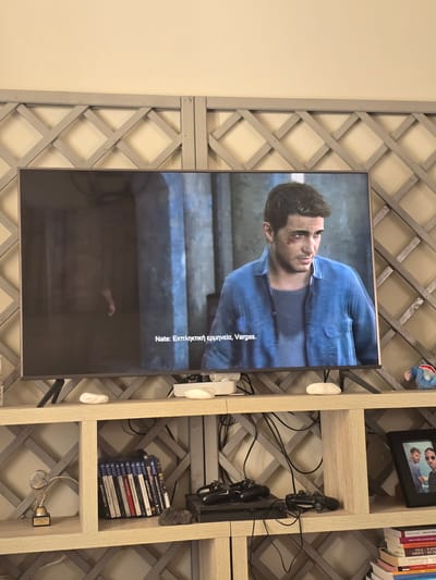 The image shows a television displaying a scene from a video game, likely "Uncharted 4," as indicated by the character on screen and the PlayStation game cases visible on the shelf below. The character, Nathan Drake, is wearing a blue shirt and has a bruised cheek. Subtitles in Greek read "Nate: Εκπληκτική ερμηνεία, Vargas." The television is placed on a light wood shelf unit with a grey lattice-patterned back. On the shelf below the TV, there are two white, smooth stones, game controllers, and a collection of "Assassin's Creed" game cases for PlayStation. To the left of the shelf is a trophy. To the right, a framed photograph shows two men, and a stack of books is visible. The environment appears to be a living room in a domestic setting. The television screen has a reflection of the room and the lattice work. The overall scene is well-lit, with the primary focus on the television and the items arranged on the shelf. The location is identified as Agii Anargiri, Greece.