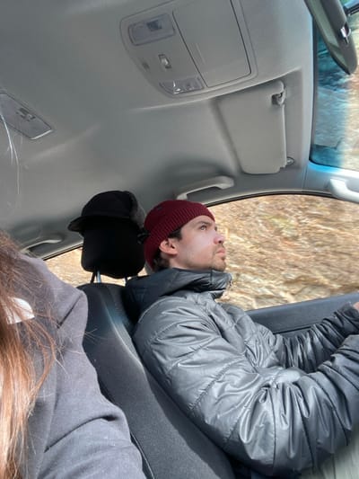 Inside a car, a person wearing a maroon beanie and gray puffer jacket looks upwards. Another person with long, brown hair sits nearby. A black hat rests on the headrest. The car's interior is light gray. The view from the window shows a blurred, grassy landscape. The scene is lit by natural light.