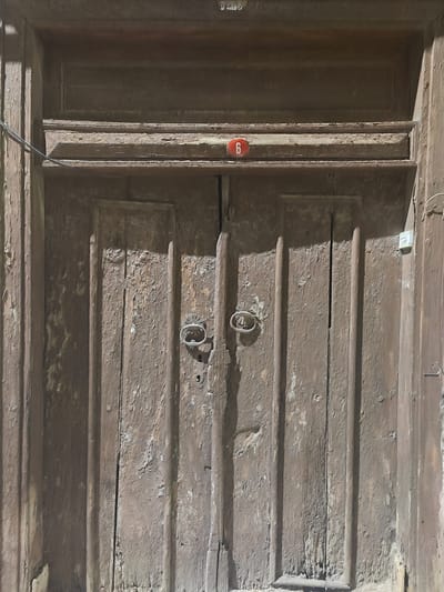 A close-up, low-angle shot captures a weathered, brown wooden double door in Osmangazi, Turkey. The door is divided vertically into two panels, each featuring raised vertical stiles. The wood exhibits significant wear and peeling paint, revealing a lighter wood underneath in many areas. Two ornate, circular metal door knockers are centrally placed on each door panel, with a visible keyhole below them. Above the door, a horizontal wooden ledge has a small, oval red sign with the white number "6" adhered to it. The top of the door is recessed, with a darker wooden frame. A thin black wire runs diagonally from the upper left corner of the image across the door's frame. A light-colored electrical outlet is mounted on the right side of the door frame. The lighting suggests an outdoor setting, with strong shadows falling across the door's surface.