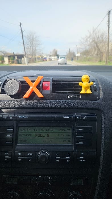 A daytime image taken from the interior of a vehicle, looking forward through the windshield. The vehicle's dark, textured dashboard occupies the lower half of the frame. Attached to the central air vents are three items: a circular black object with white markings on the left, a bright orange 'X'-shaped object in the center, and a small yellow smiling humanoid figure on the right. A red triangular hazard light button is visible between the orange 'X' and the yellow figure. Below the dashboard items, a SkodaAuto brand car radio and infotainment system displays green text, including "FM-AS 13:10", "KANAL 77", "FOOL'S", "18.5 °C", and "87.50". Additional climate controls below the radio show "OFF".

Through the windshield, a paved road stretches into the distance under a clear, light blue sky. A white SUV is observed driving away from the viewer in the mid-distance. On the right side of the road, a solitary individual, appearing as a blurred figure, walks away from the camera. The roadside environment includes bare deciduous trees and several wooden utility poles with overhead wires. Low-rise buildings are visible along the left side of the road in the background. This scene is located in the city of Gevgelija, North Macedonia.